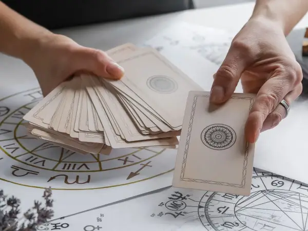 Dłonie trzymają talię kart, jedna karta jest wyciągnięta. W tle widać diagramy astrologiczne. To zapowiedź wróżby z **tarot 4 karty**.